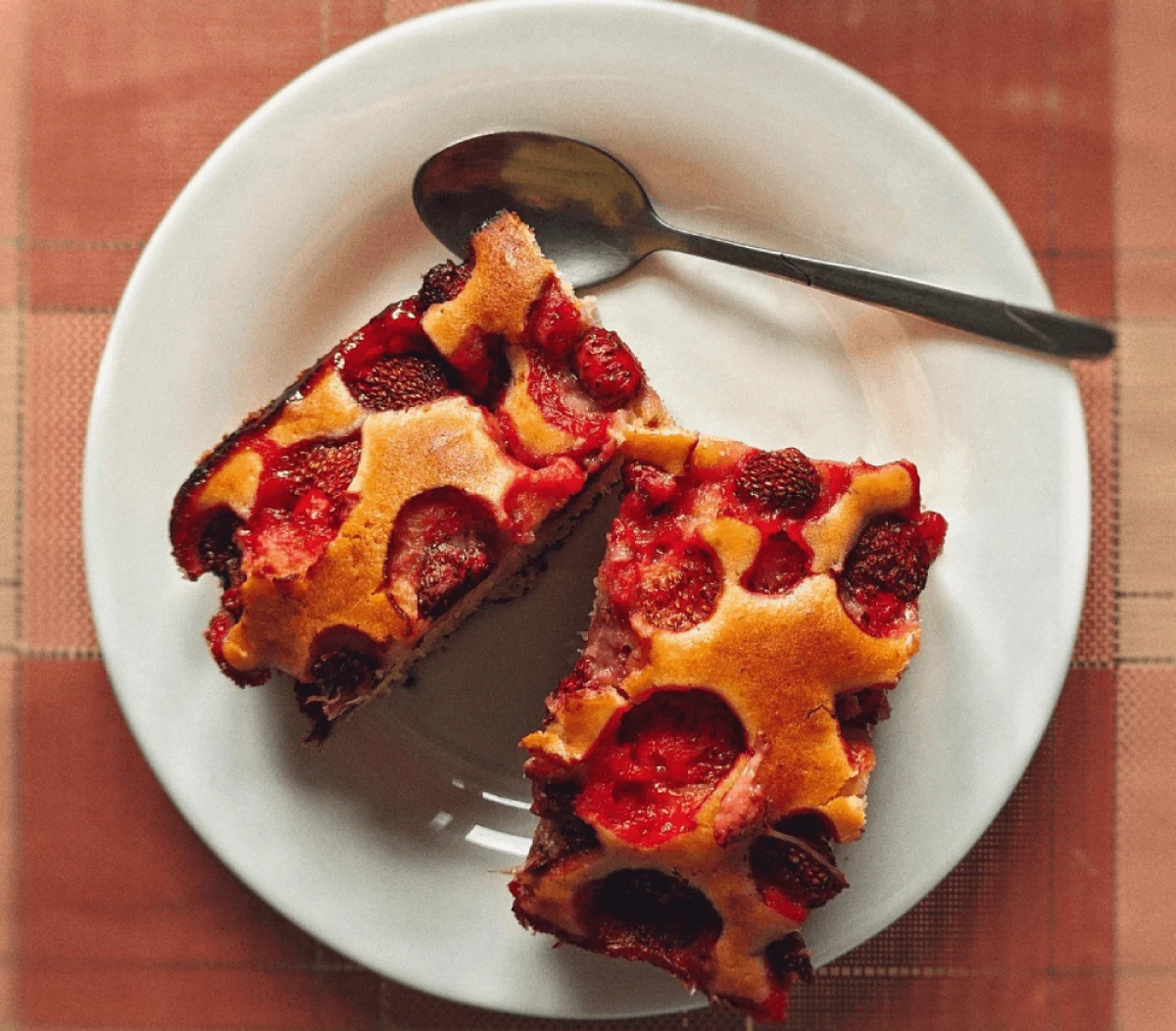 Na białym talerzu leżą dwa kawałki ciasta z truskawkami, obok leży łyżeczka